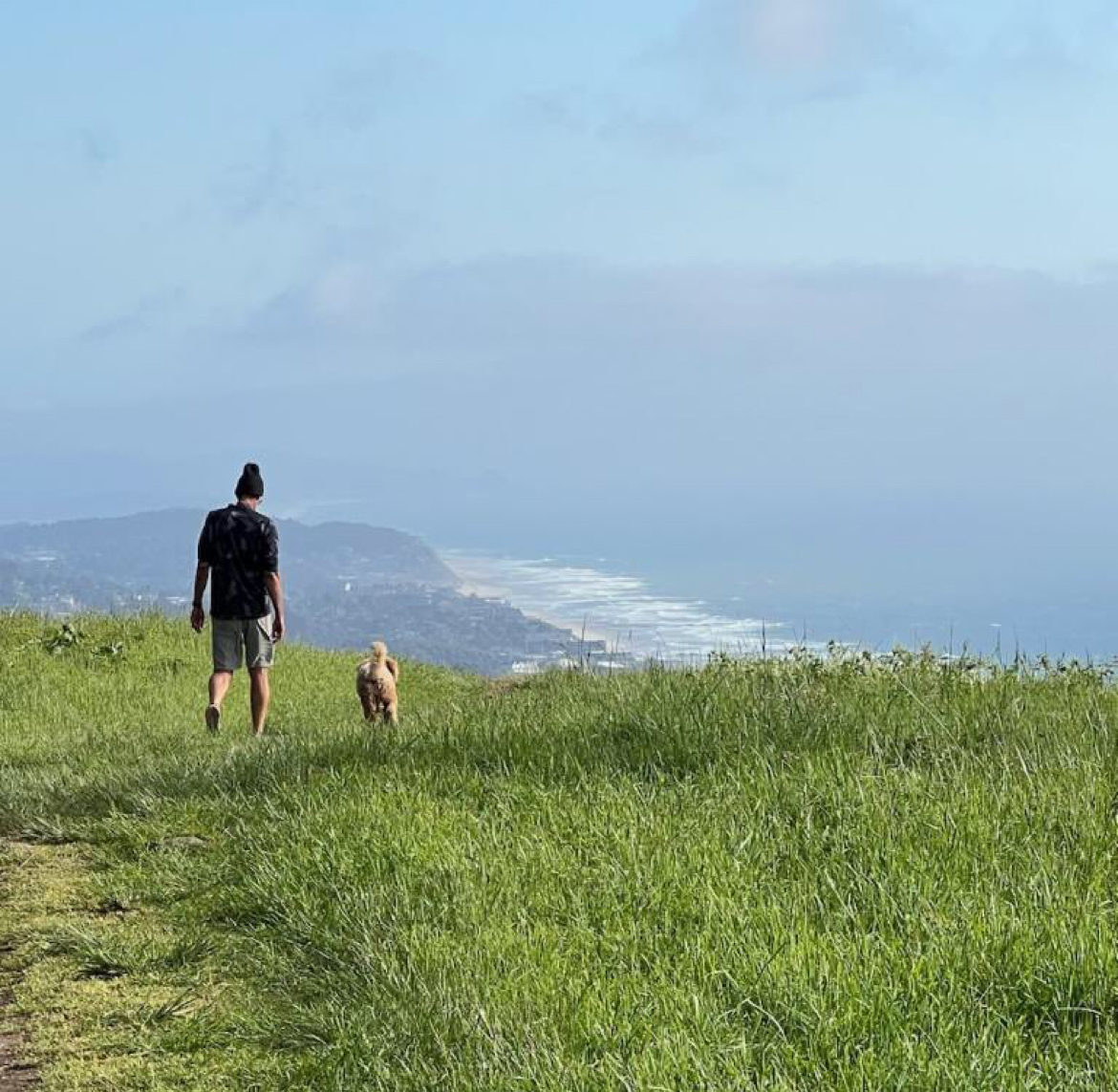 Man In Black Hat And T-shirt Walking With Dog On Green Grass